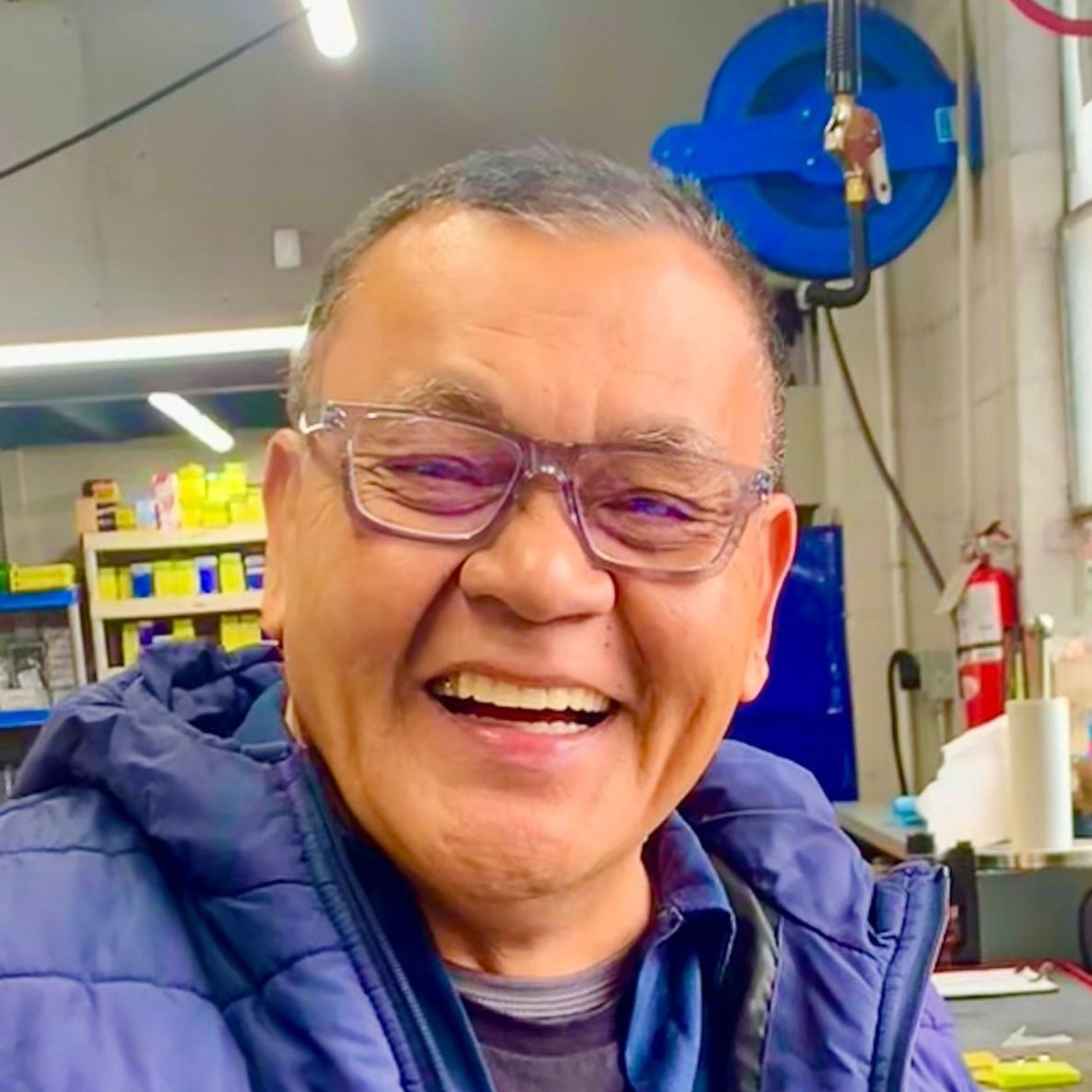 Man wearing glasses smiles broadly in a workshop, wearing a blue jacket.