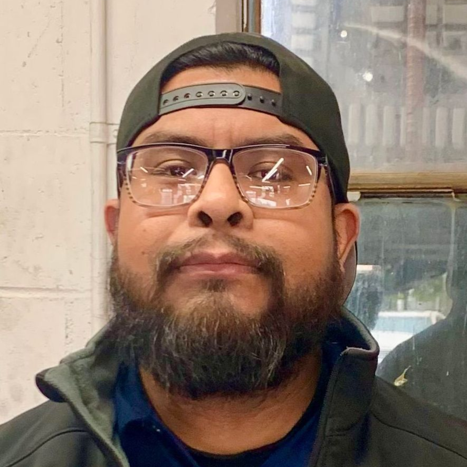 Man wearing glasses, a baseball cap, and a blue shirt, in front of a window.