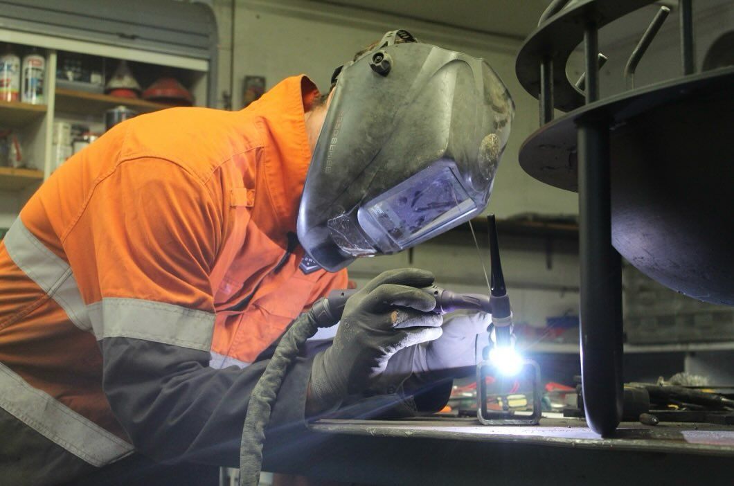 Welder in orange and navy workwear using a welding torch, inside a workshop.