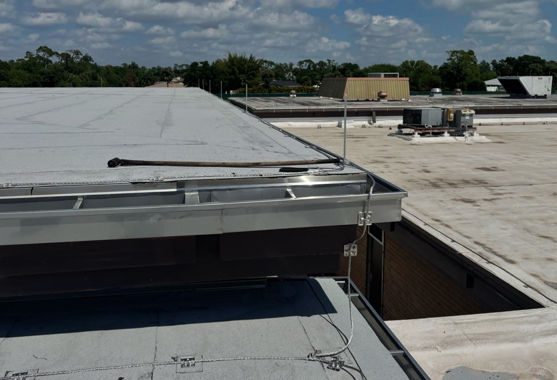 A rooftop view showing a metal gutter system, roofing material, and a lightning rod on a clear, sunny day.