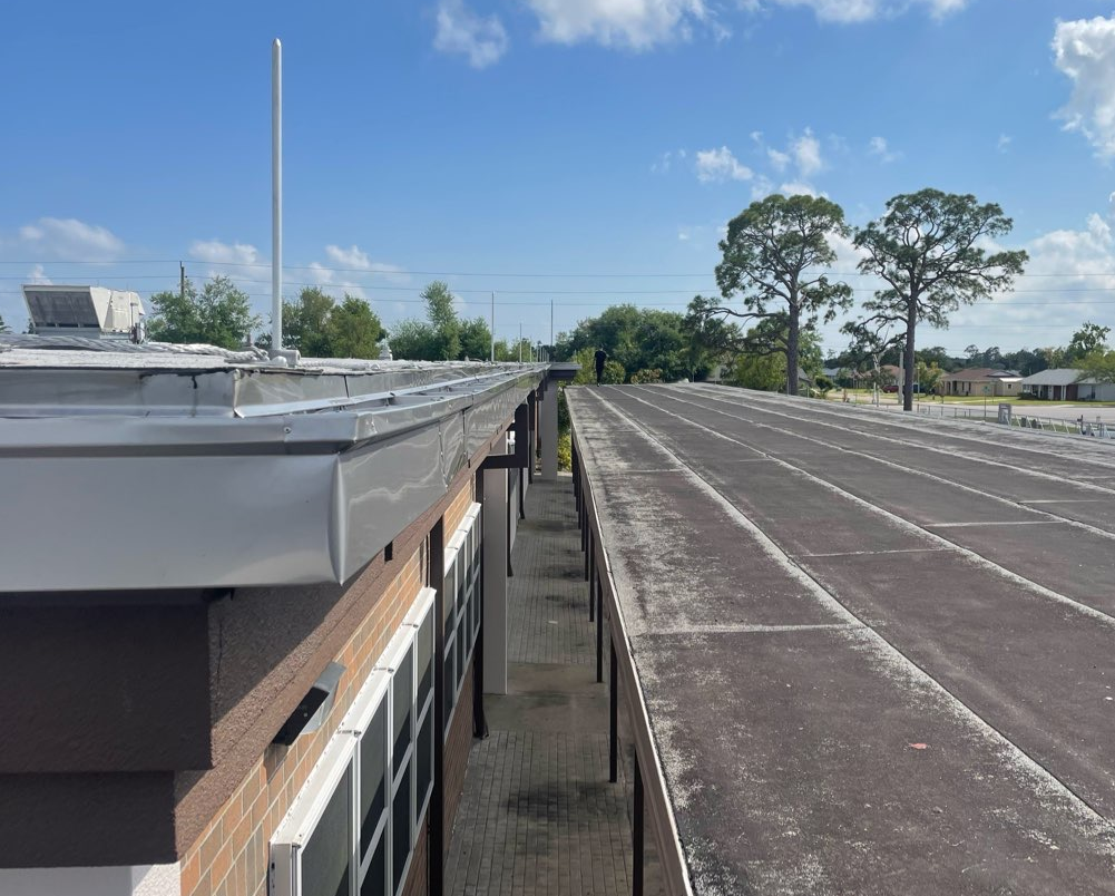 A rooftop view shows a modern building exterior with a flat, grey roof alongside a bright metal-trimmed section under sky.