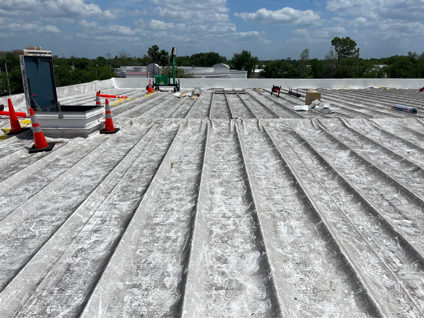 A flat industrial roof undergoing maintenance with a roof hatch, orange safety cones, and a lift in the background.