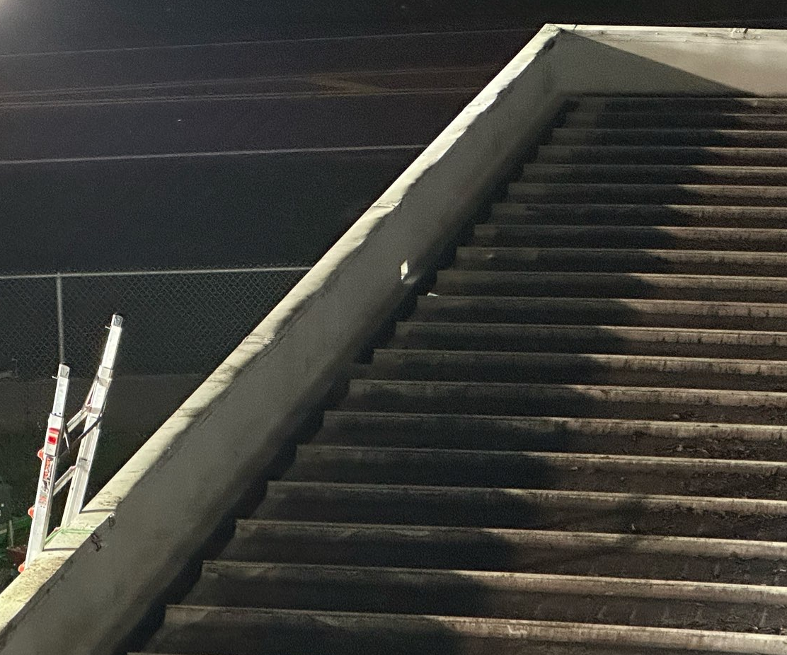 A set of concrete outdoor stairs at night with a metal ladder leaning against a wire fence to the left.