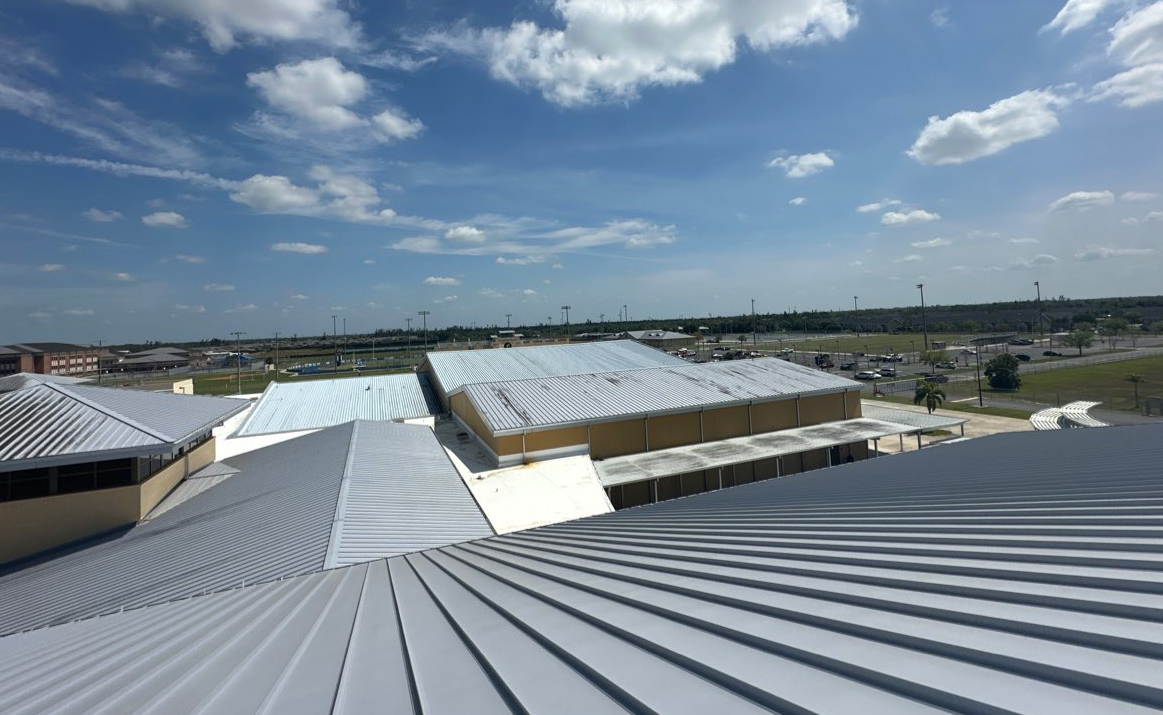 A high-angle view of multiple gray metal roofs on a commercial building under a bright blue, cloudy sky.