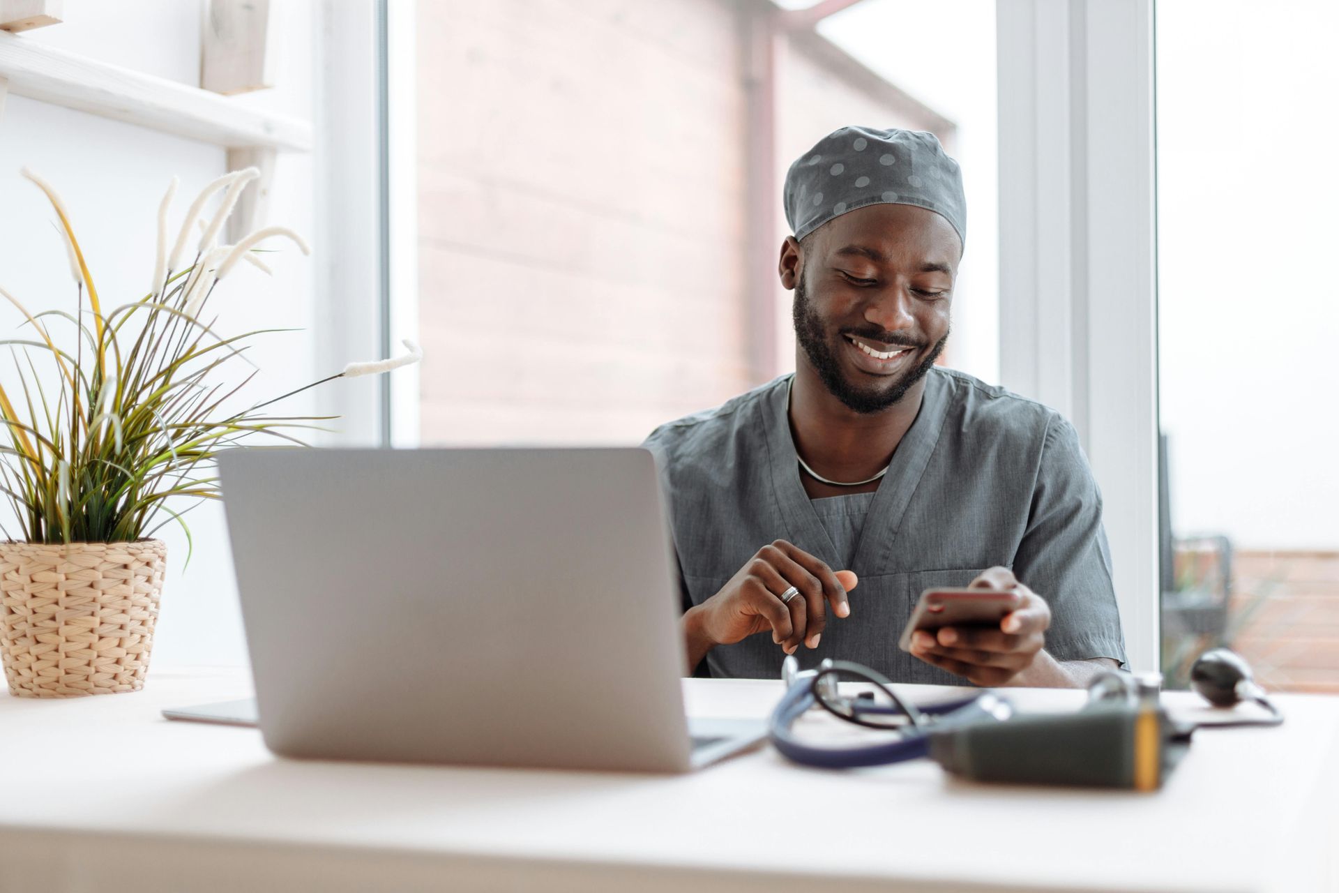 Man in scrubs smiles at phone, seated at desk with laptop, stethoscope, and plant.