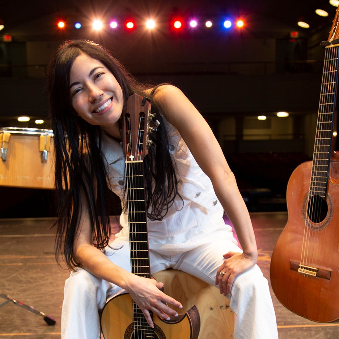 Smiling woman sitting on a cajón on a stage with guitars and colored lights behind her