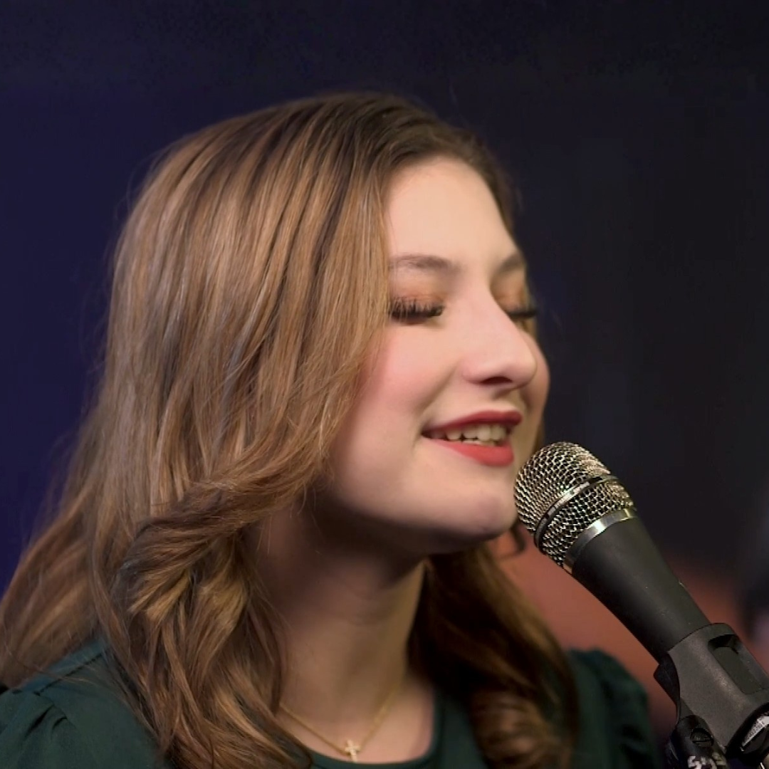 Woman singing into a microphone, eyes closed, in a studio with a dark background.