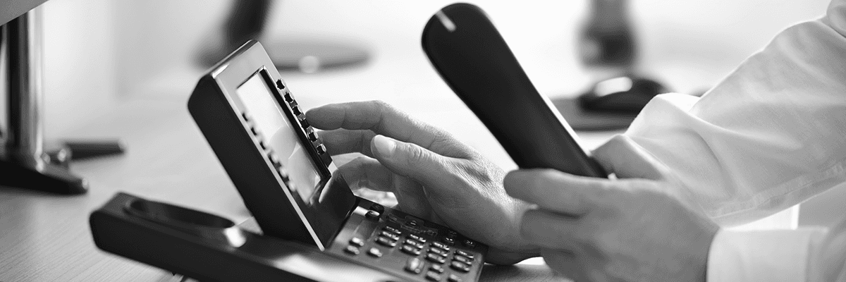 Eine Person benutzt ein schwarzes Bürotelefon und hält den Hörer in der Hand. Das Telefon steht auf einem Schreibtisch.