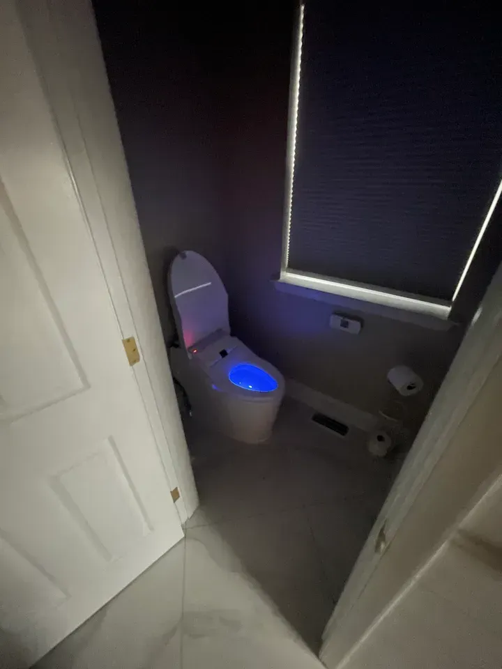A modern toilet with blue and white lights in a small bathroom with a closed door and a window.