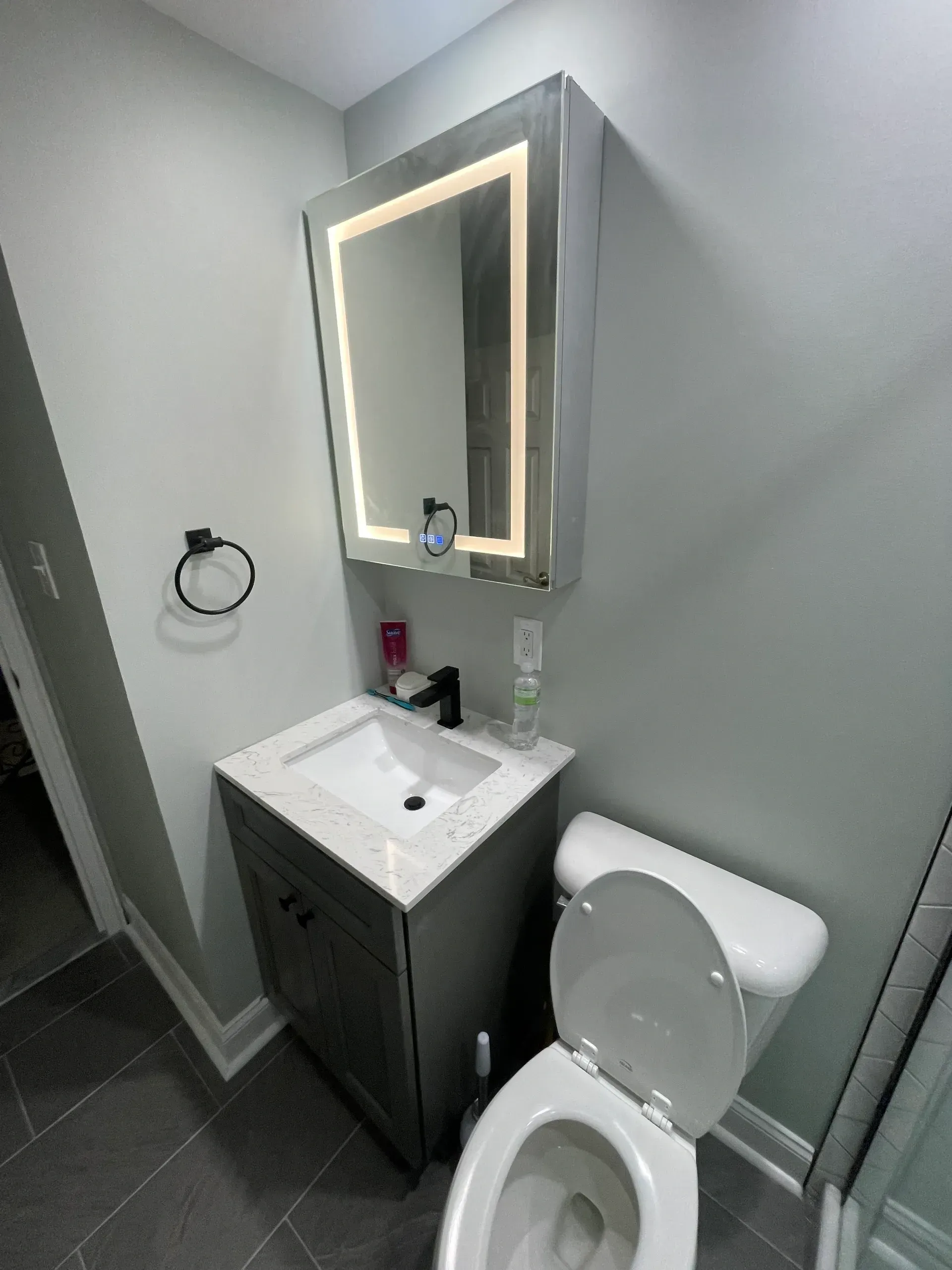 Small gray bathroom with a toilet, sink, and lighted mirror.
