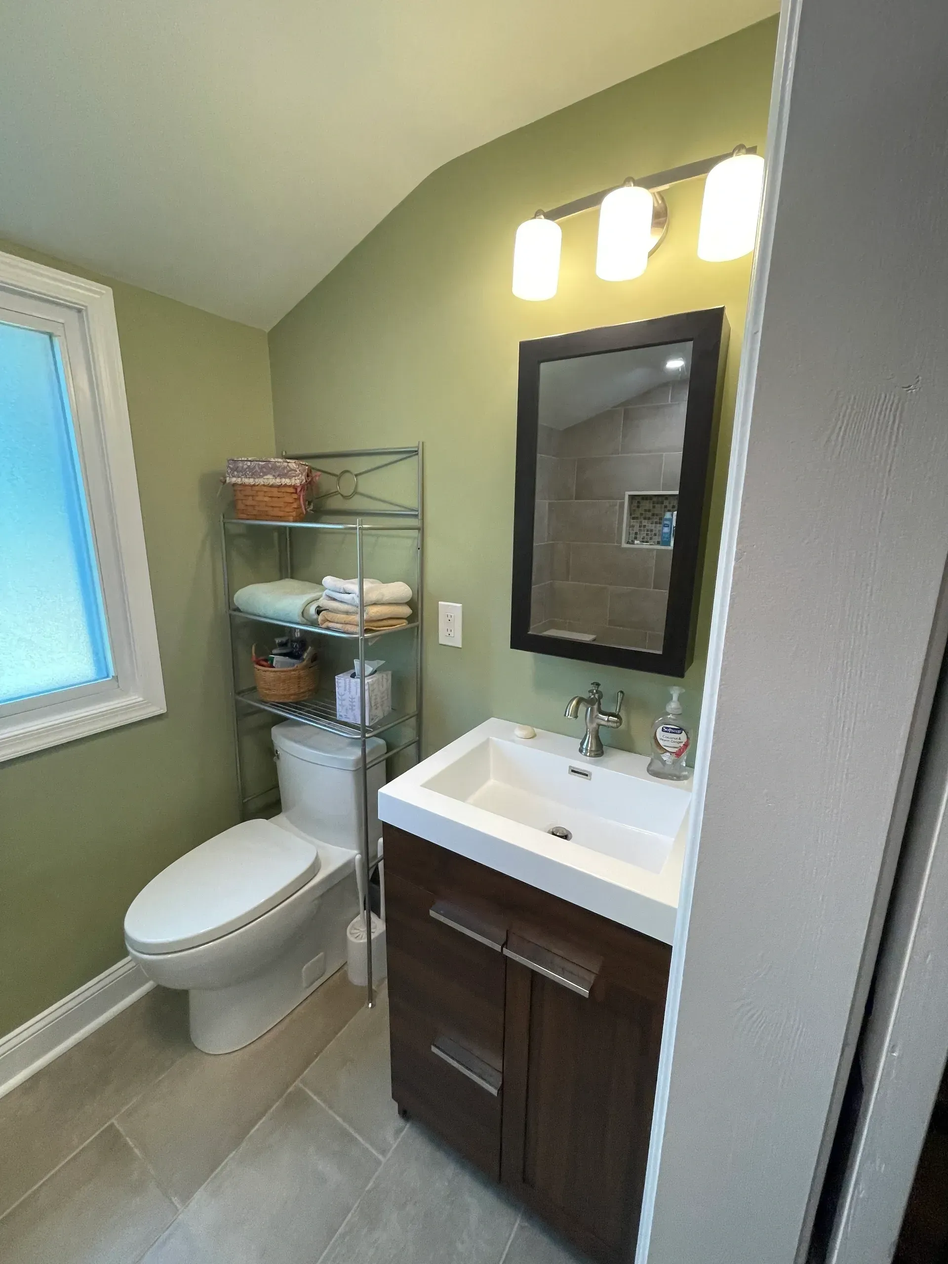 Bathroom with green walls, a white toilet, brown vanity, and a metal shelving unit.