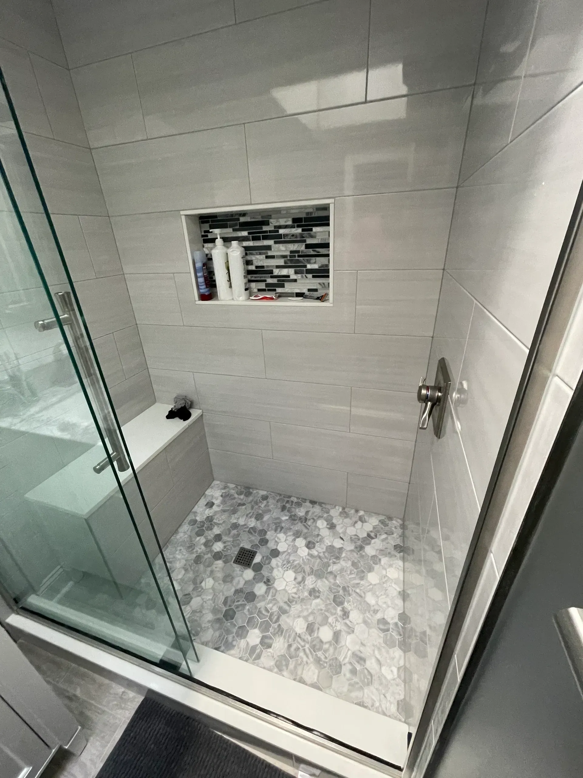 Modern shower with gray tiled walls, pebble floor, built-in bench, and glass door.