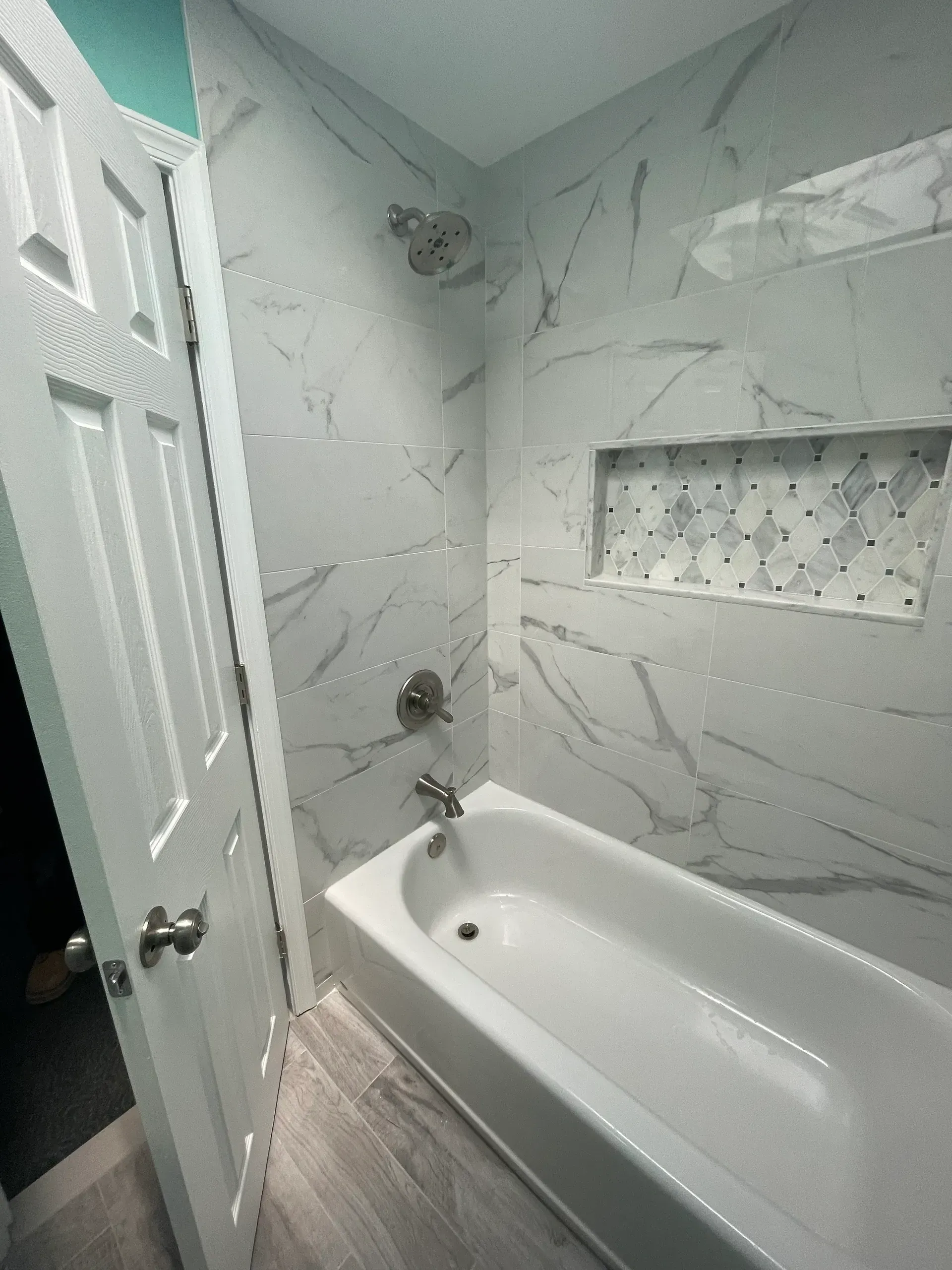 White bathroom with marble-like tile, a built-in shelf, and a white bathtub. 