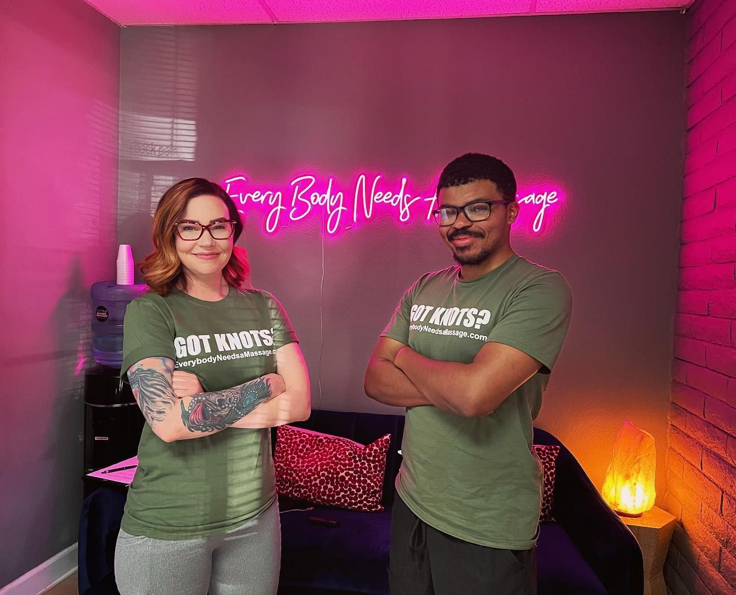 Two people stand with arms crossed in a room; neon sign reads