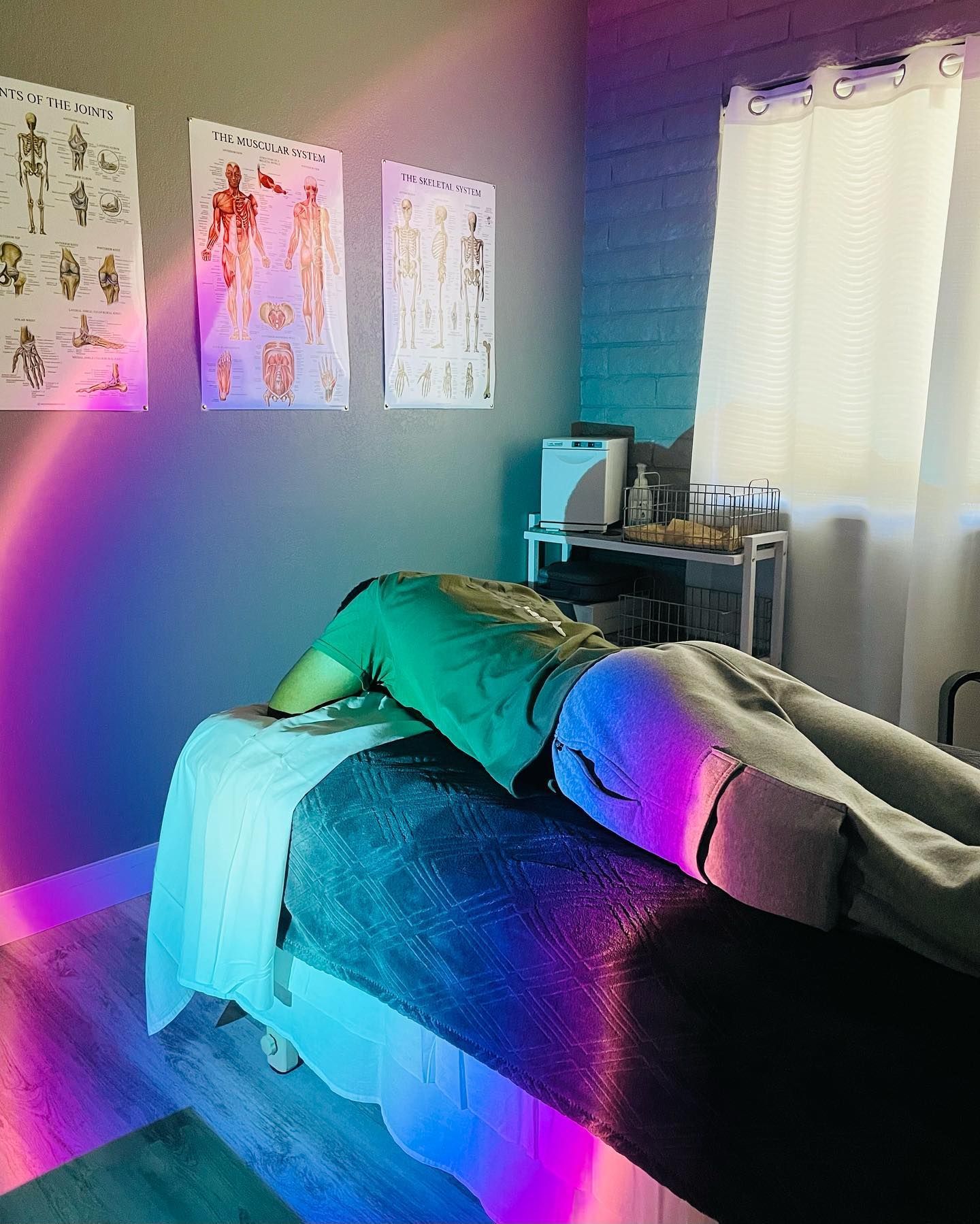 Person lying face down on a massage table, in a colorful room. Medical posters on wall.
