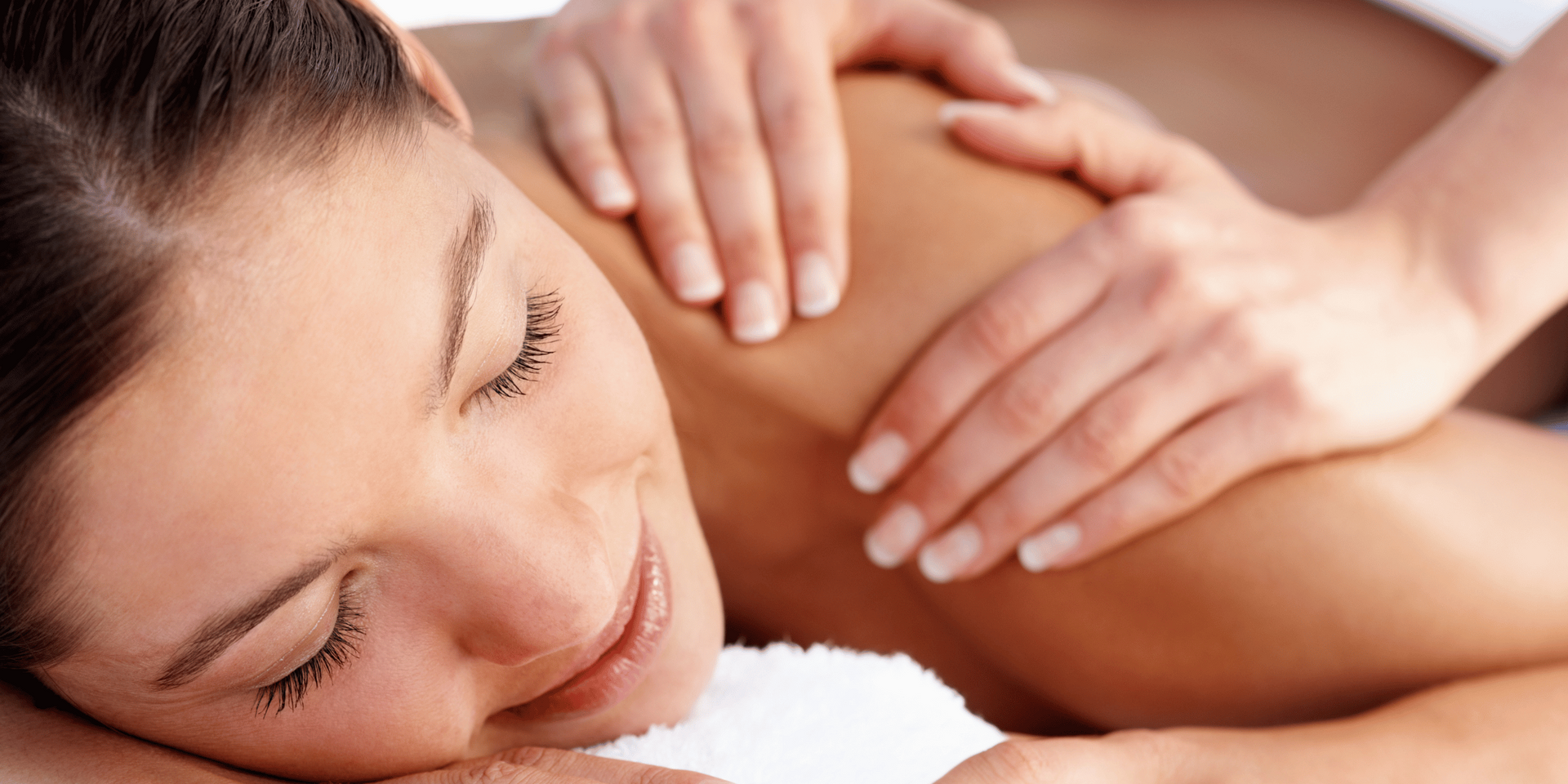A person receiving a shoulder massage while lying on a white towel, eyes closed, smiling.