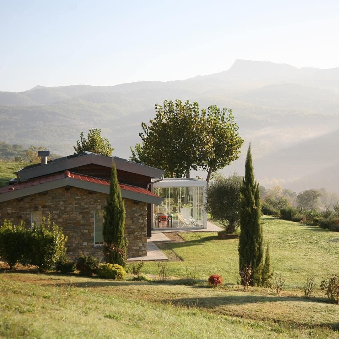 una piccola casa in pietra si trova in mezzo a un prato verde con le montagne sullo sfondo .