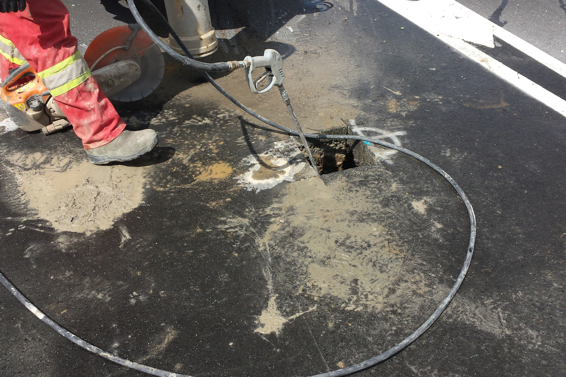 Pessoa de macacão vermelho corta asfalto na estrada, criando um buraco quadrado perto da linha branca.