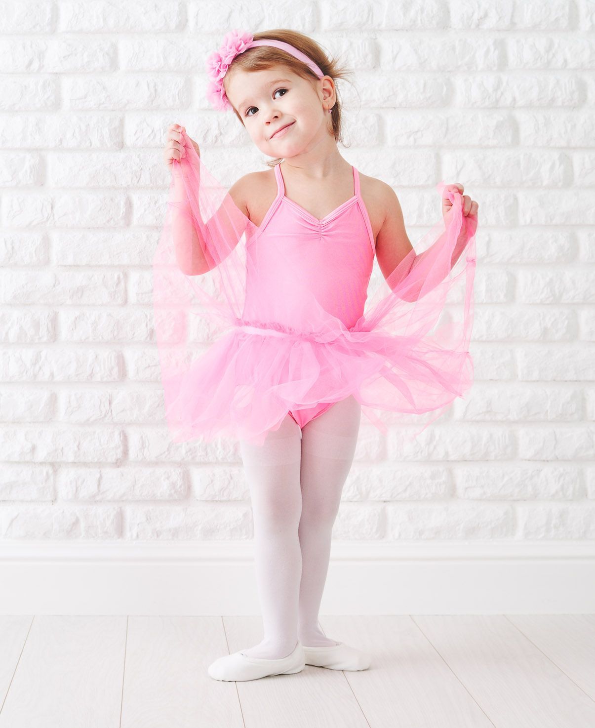 A little girl in a pink tutu and gloves is standing in front of a white brick wall.