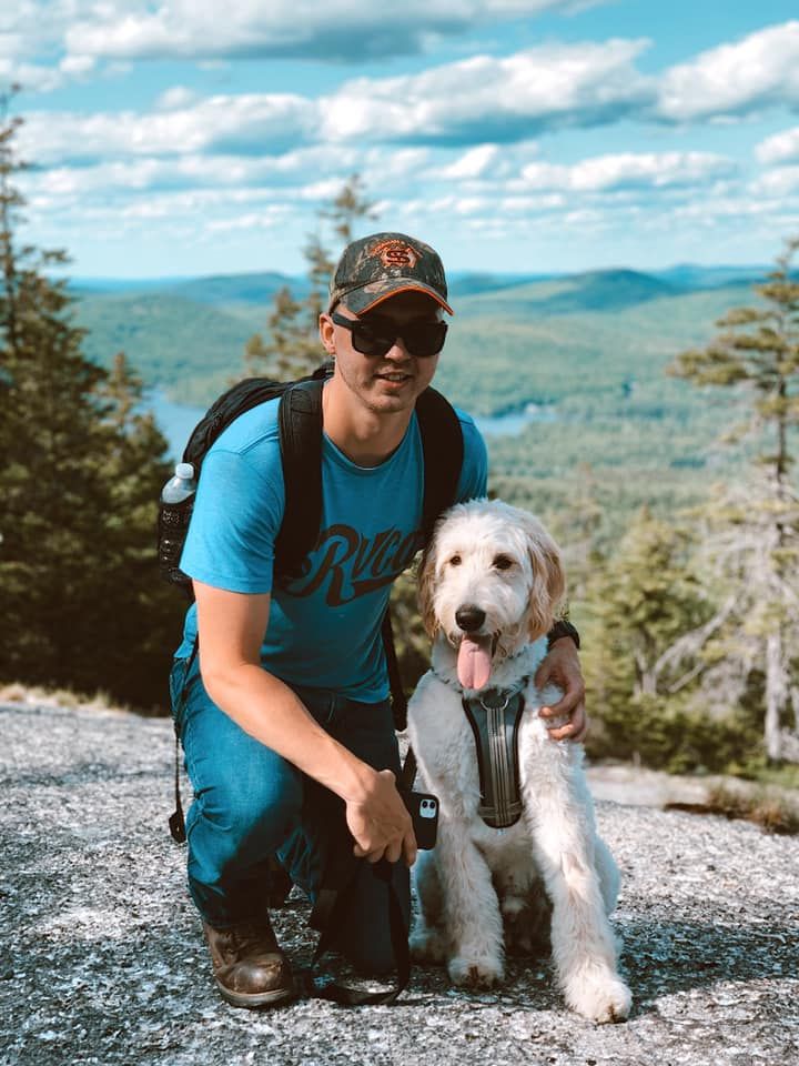 Man and dog on a mountaintop. Man kneels, petting a golden doodle, scenic view in background.