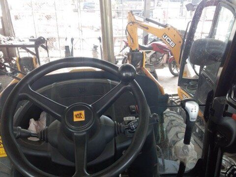 Interior view of a JCB backhoe, showing the steering wheel and controls. Another backhoe and motorcycle are visible outside.