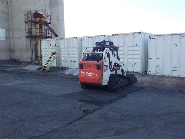 Bobcat skid-steer in a paved area, with shipping containers and a grain silo in the background.