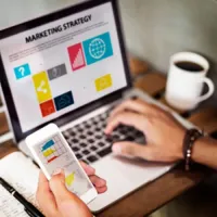 Hand using smartphone beside laptop showing a marketing strategy presentation, with coffee cup on desk