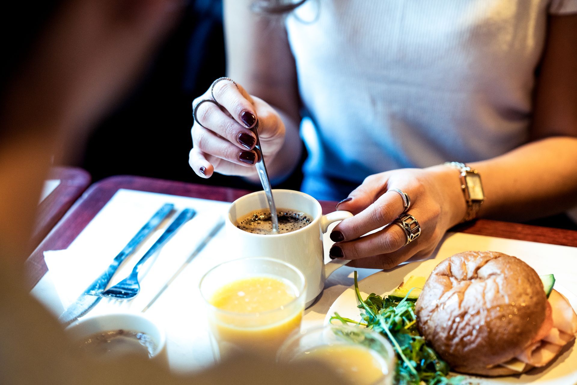 Person stirring a cup of coffee at a breakfast table with a croissant, juice, and utensils