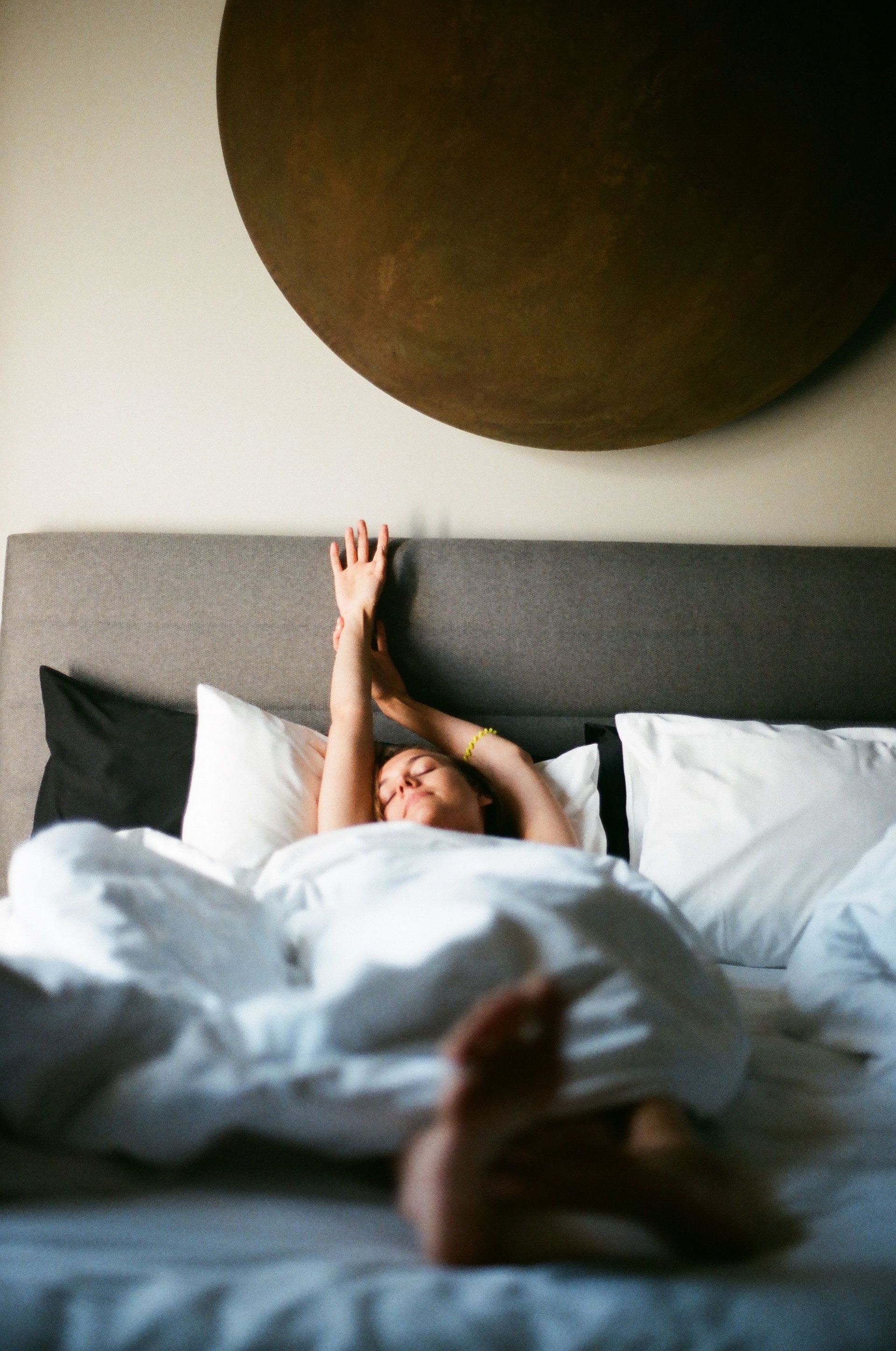 Person stretching in bed under white covers in a gray bedroom with a large round wall hanging.