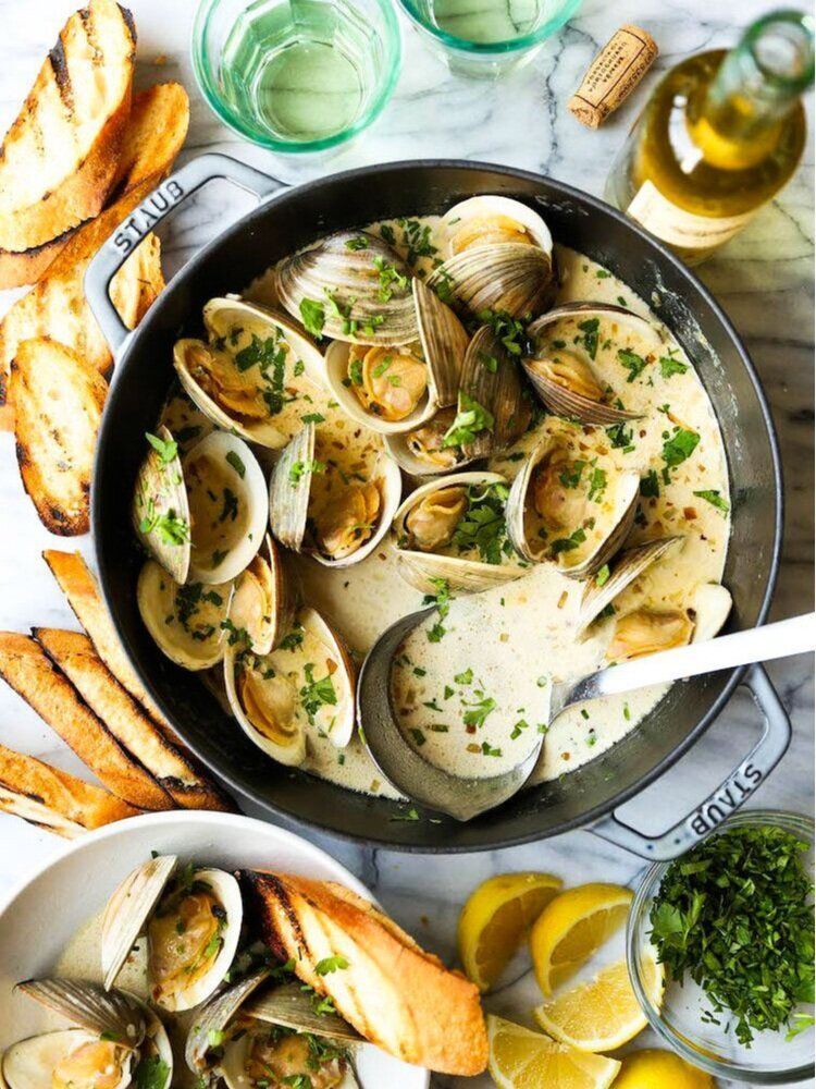 Clams in garlic herb broth with bread, lemon wedges, and wine on a rustic table