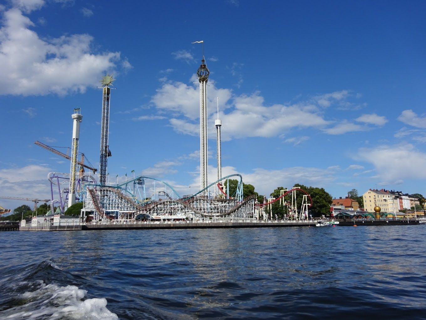 Amusement park waterfront with tall rides and roller coasters under a blue sky, seen across the water