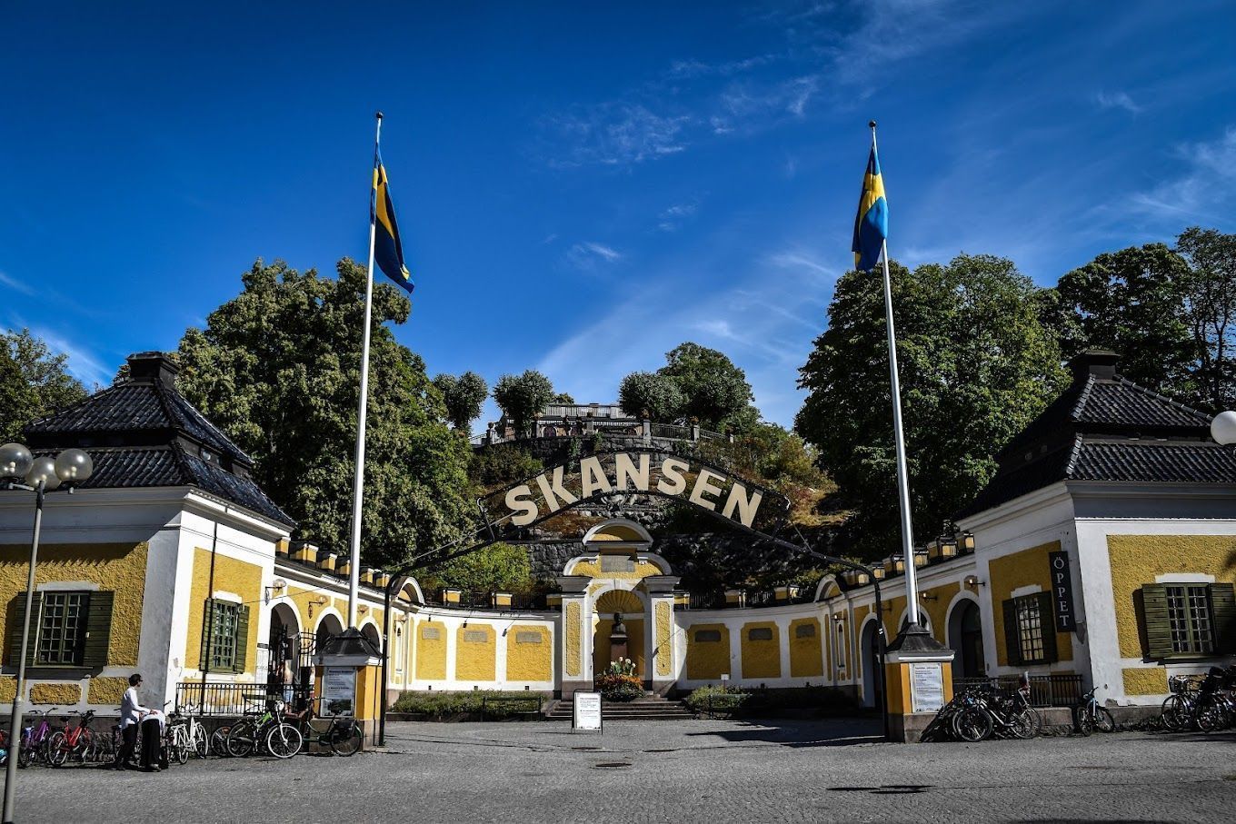 Skansen entrance gate with yellow buildings, flagpoles, and a blue sky backdrop