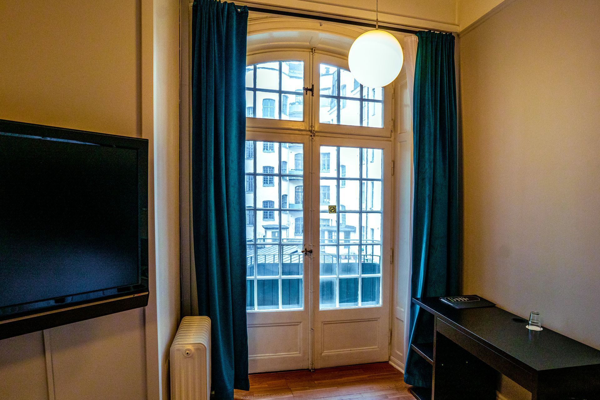 Small room with a wall-mounted TV, windowed balcony doors, blue curtains, and a black desk.