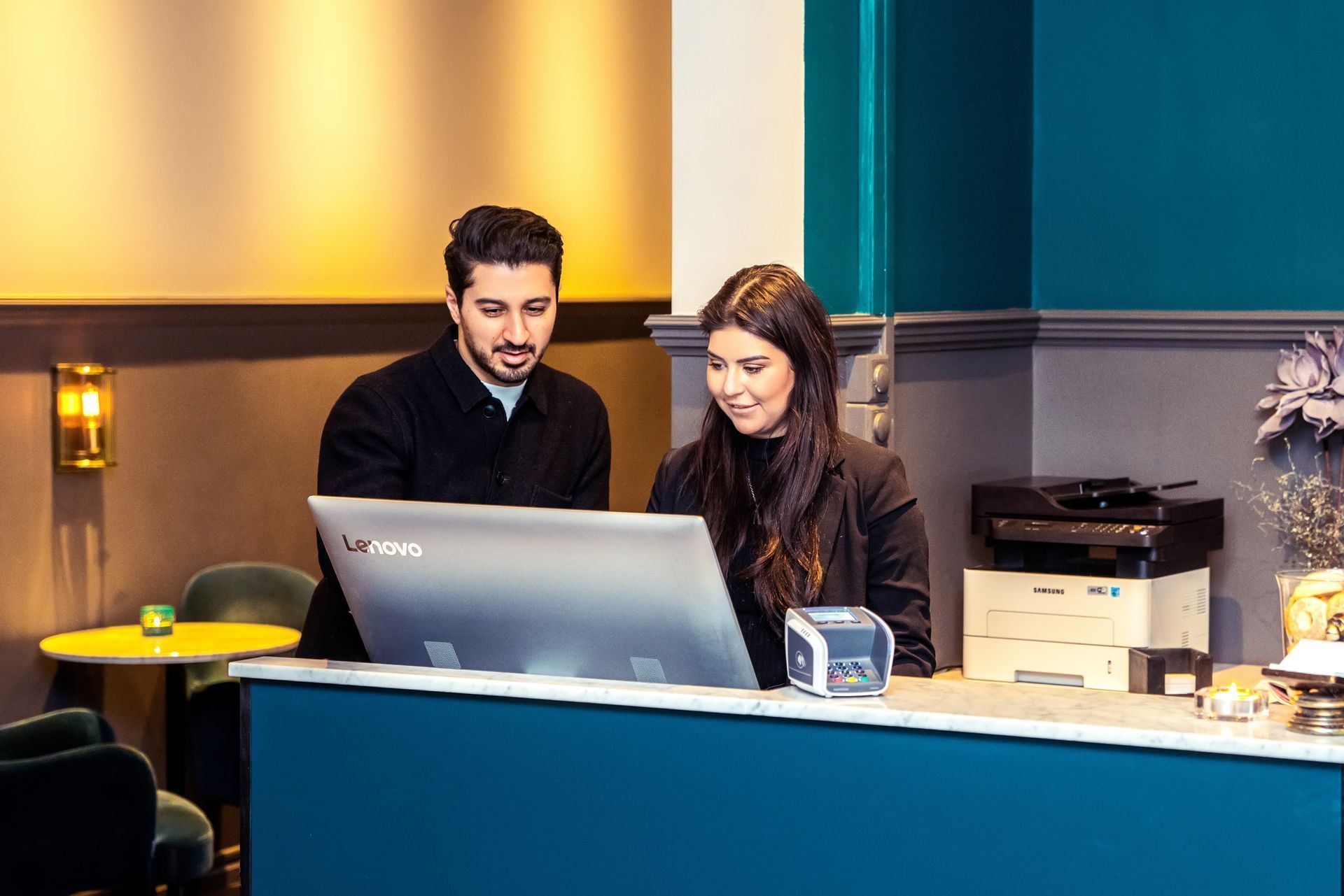 Two people standing at a reception desk, looking at a laptop in a modern office lobby.