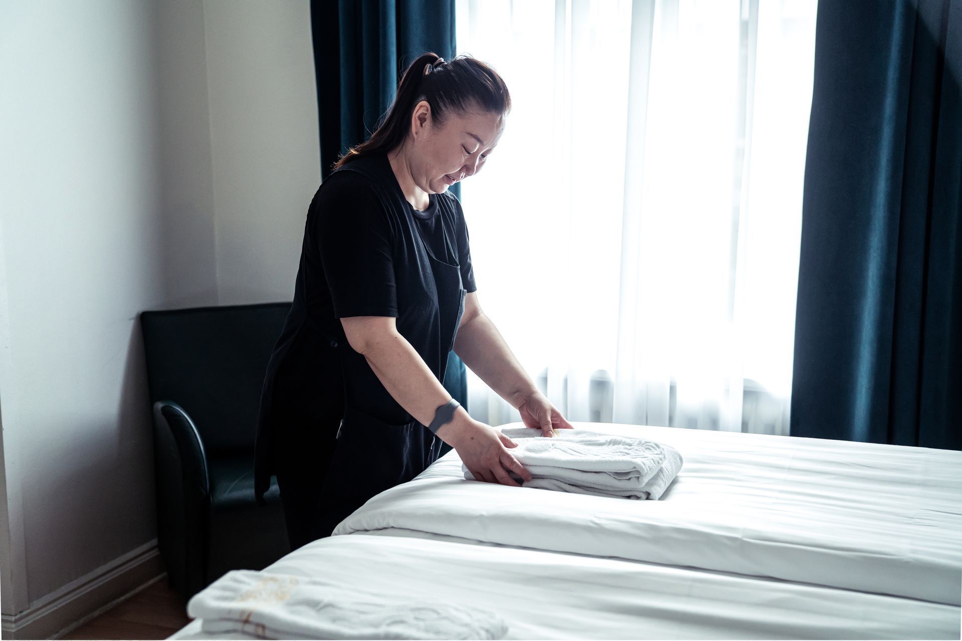 Person making a bed in a bright room with white bedding and dark curtains