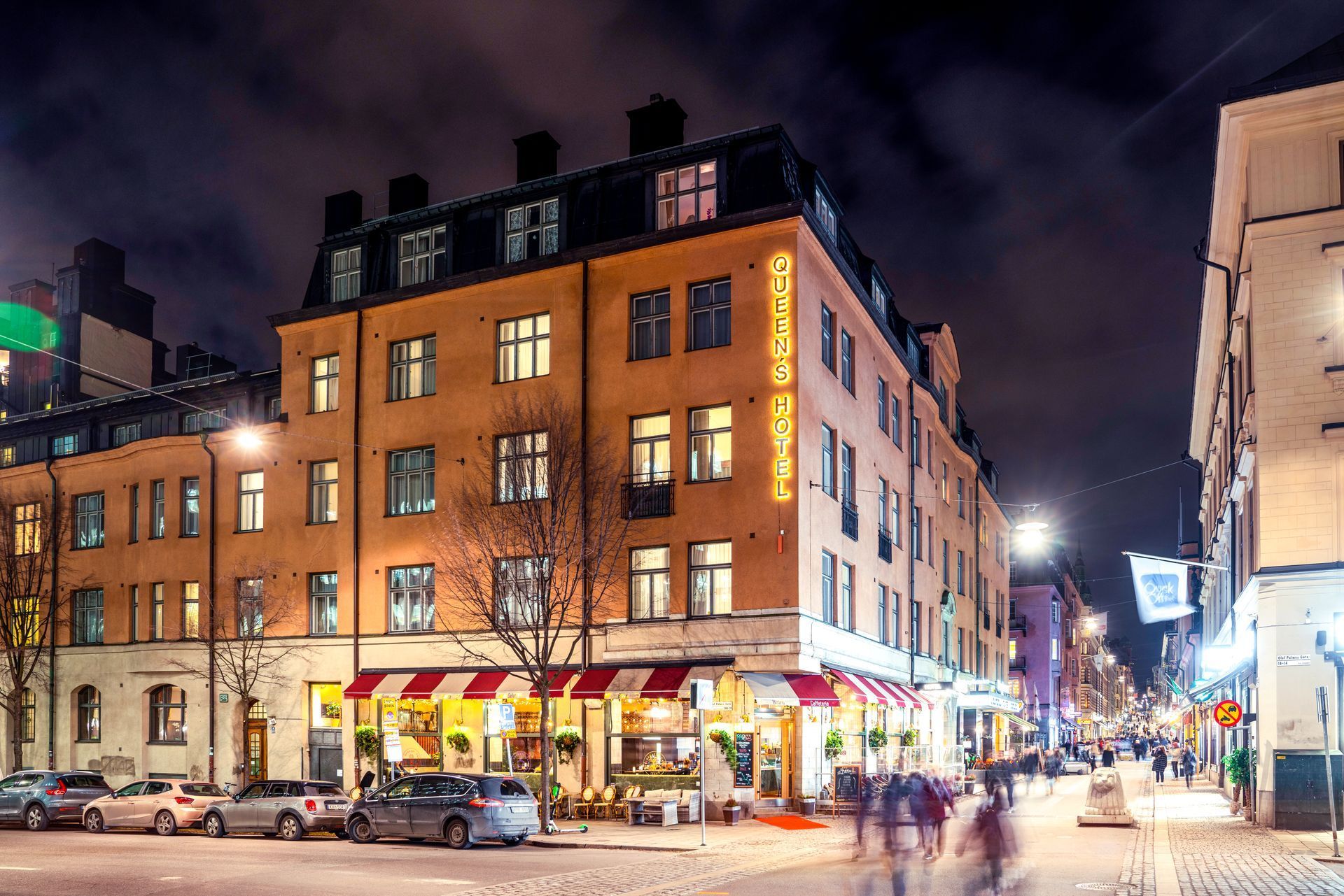 Nighttime city street with a brick corner building, lit storefronts, and pedestrians under a cloudy sky