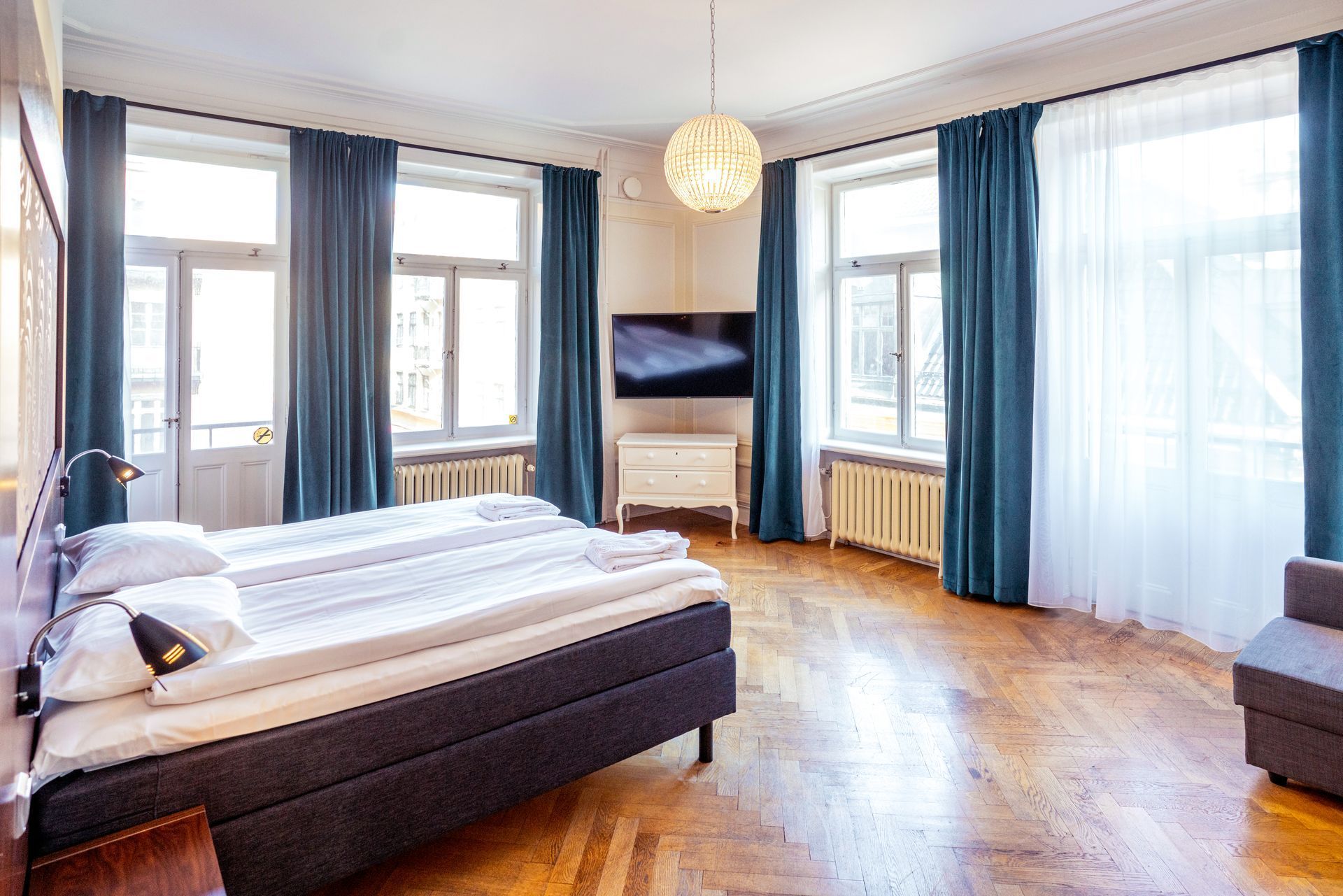 Bright bedroom with large windows, teal curtains, bed, wall-mounted TV, and hardwood floors