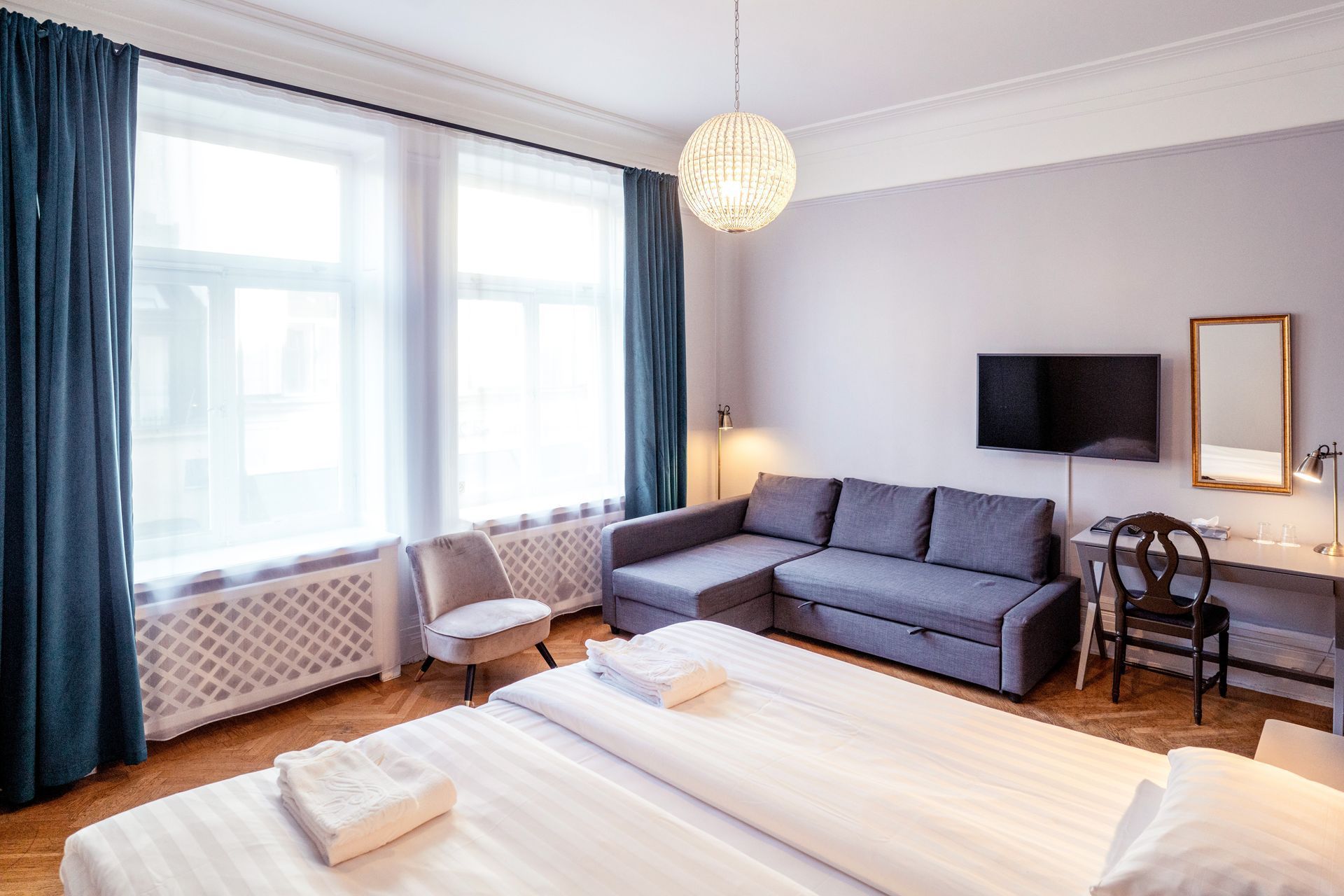 Bright bedroom with two beds, a gray sofa, wall-mounted TV, and large windows with blue curtains.