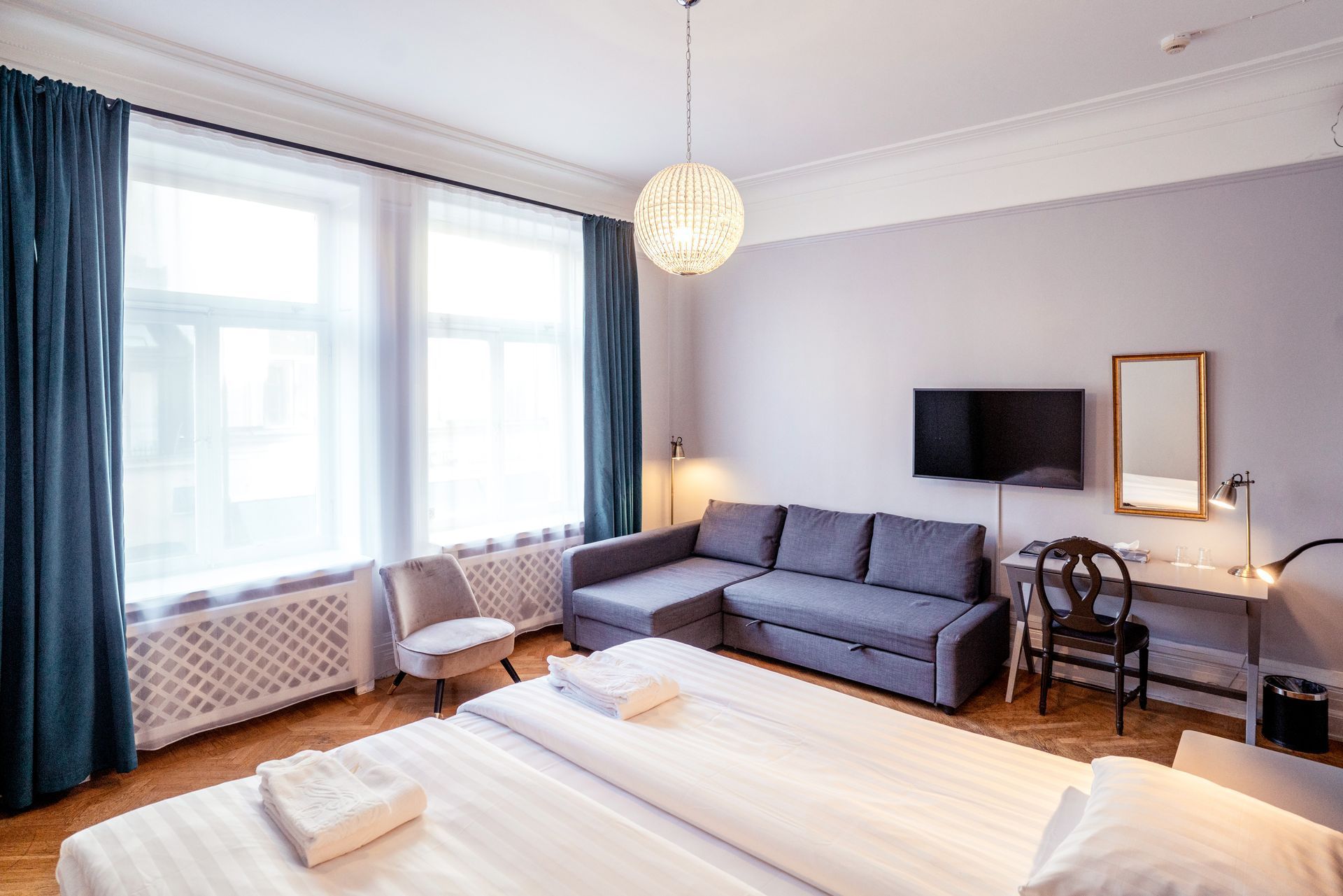 Bright modern living room with gray sofa, wall-mounted TV, large windows, and a bed in the foreground