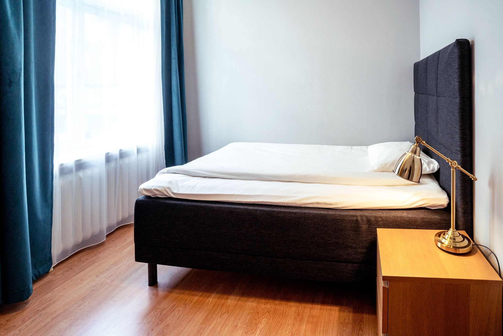 Minimal bedroom with a black bed, blue curtains, wooden floor, and a small bedside lamp