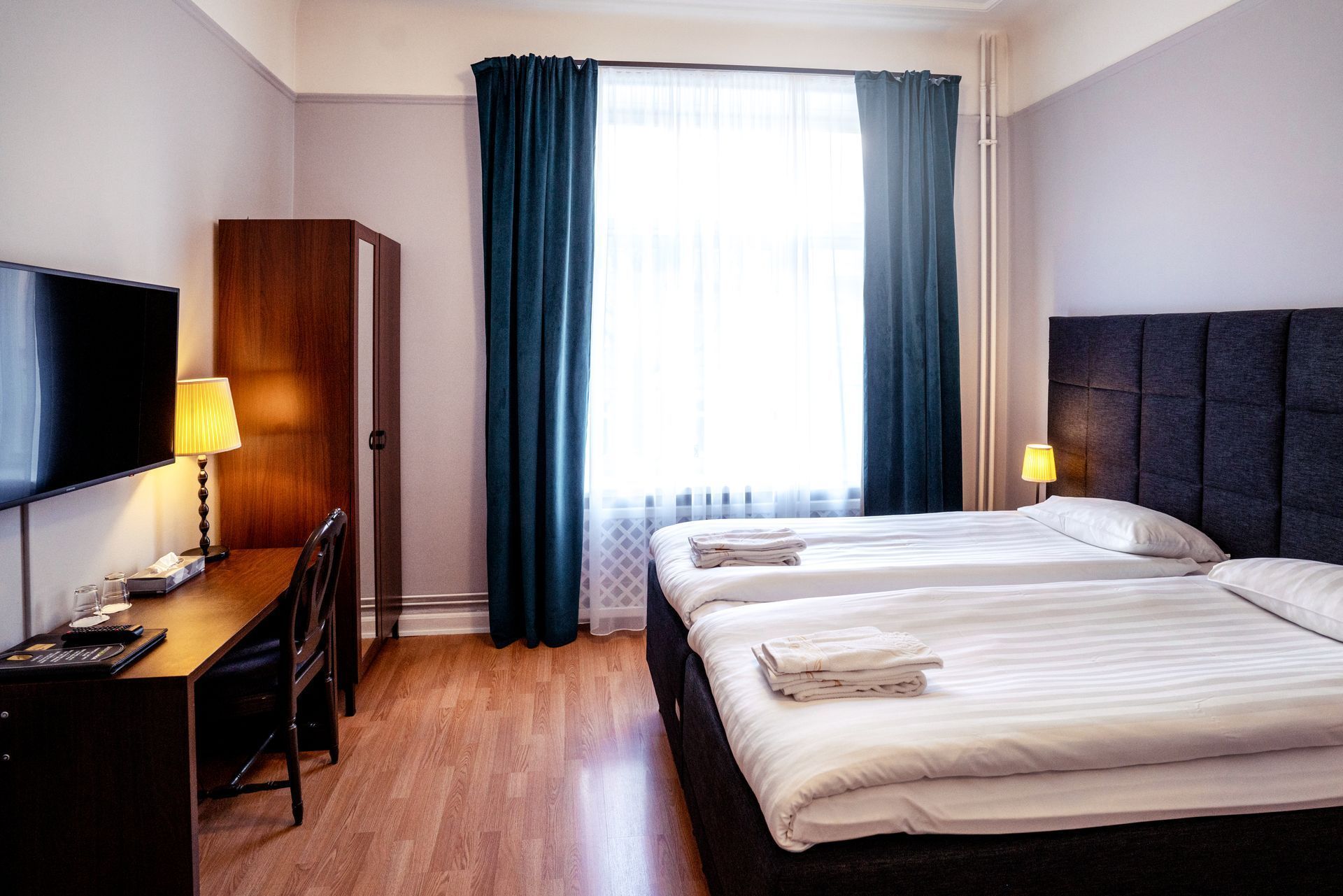 Modern hotel room with two twin beds, desk, wardrobe, and large window with blue curtains