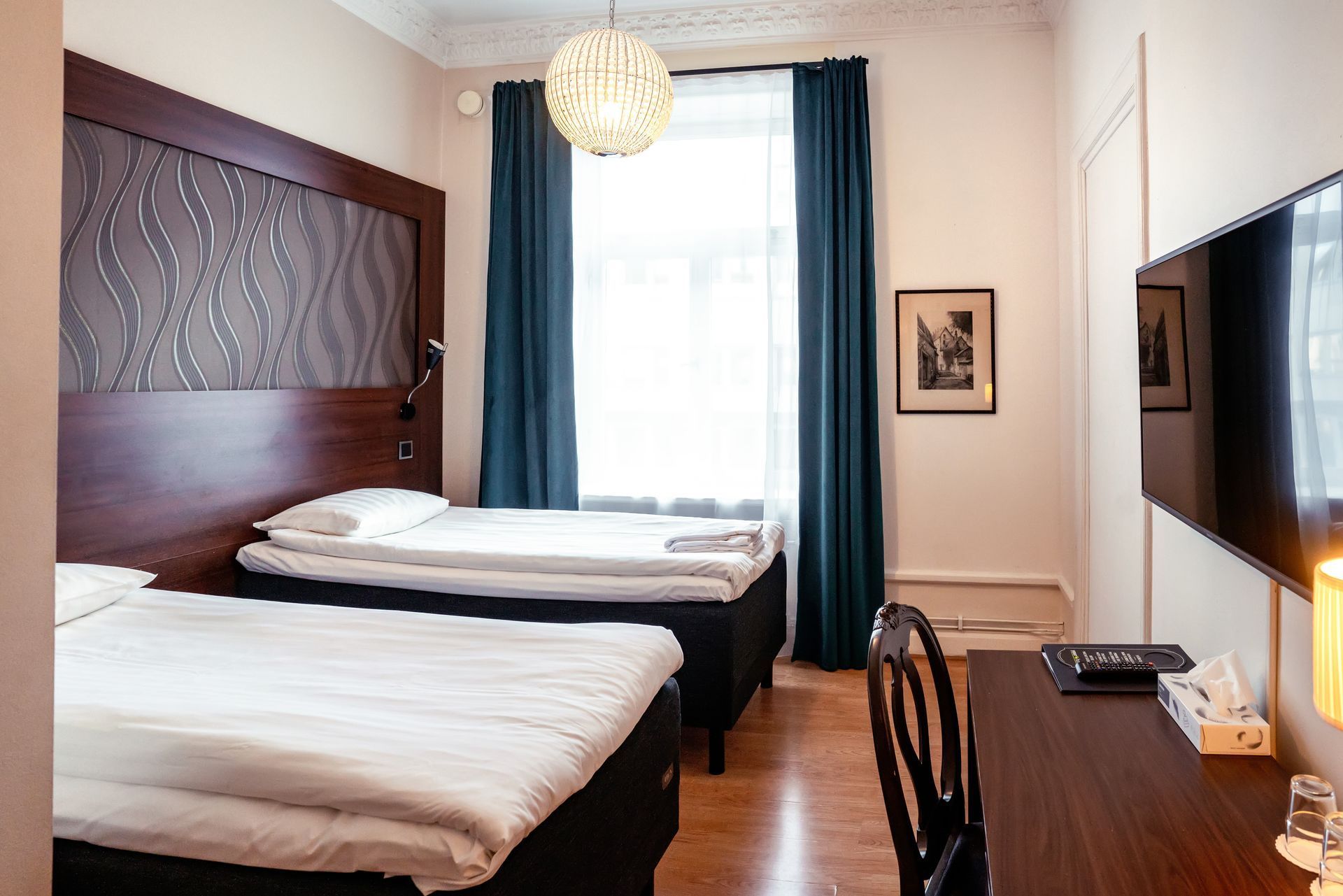 Modern hotel room with two beds, dark curtains, desk, wall art, and a TV.