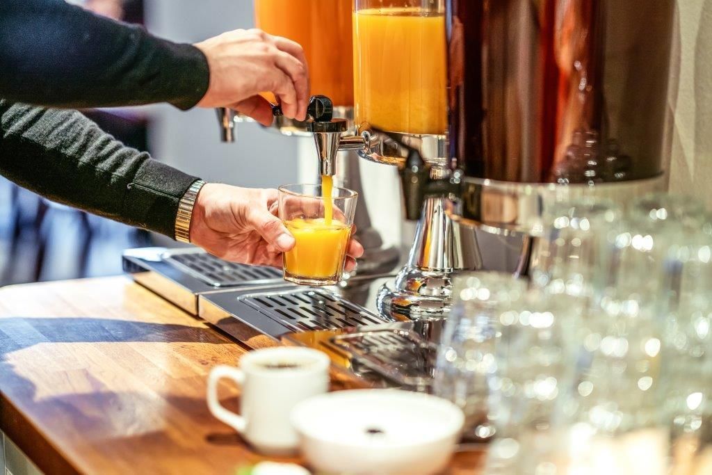 Hands pouring orange juice at a buffet counter with cups and glasses nearby