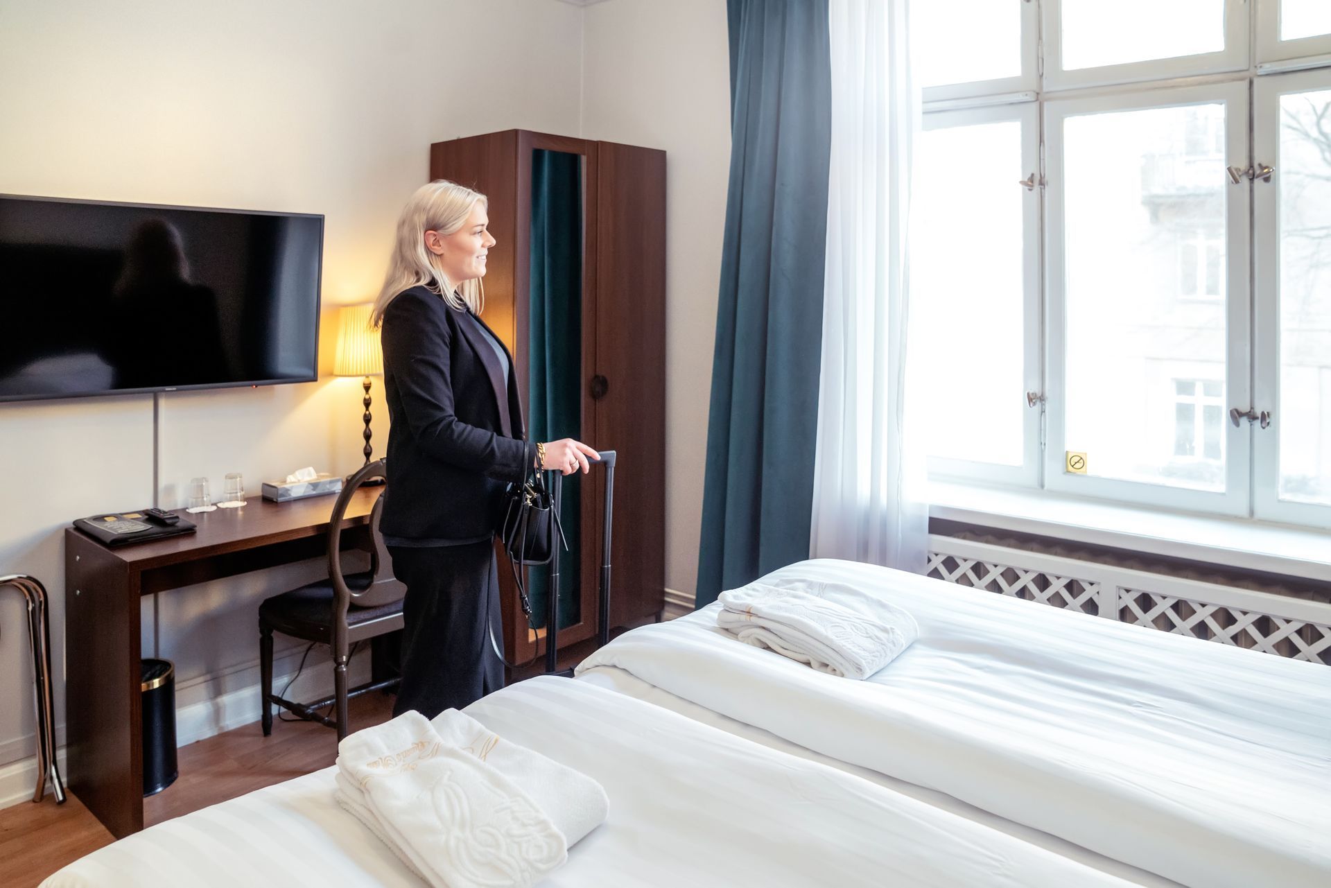 Person standing by a bed in a bright hotel room, near a window and TV.