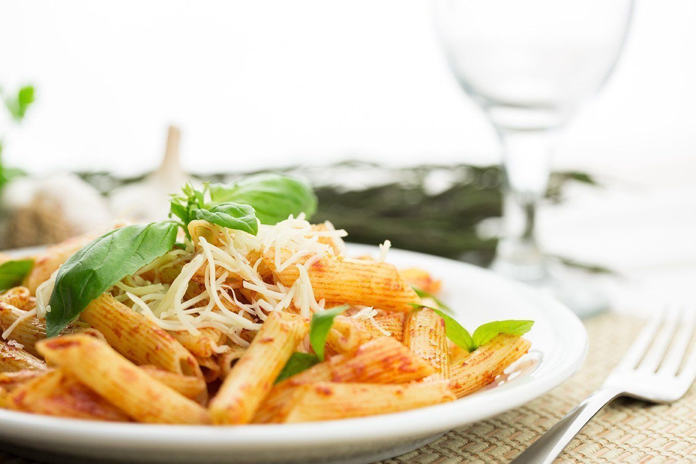 Plate of penne pasta with tomato sauce, basil, and grated cheese on a table with a wine glass in the background