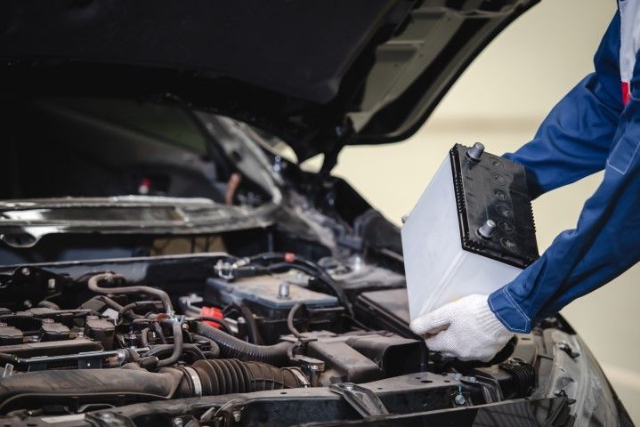 Mechanic in blue coveralls replacing a car battery in an open engine bay.