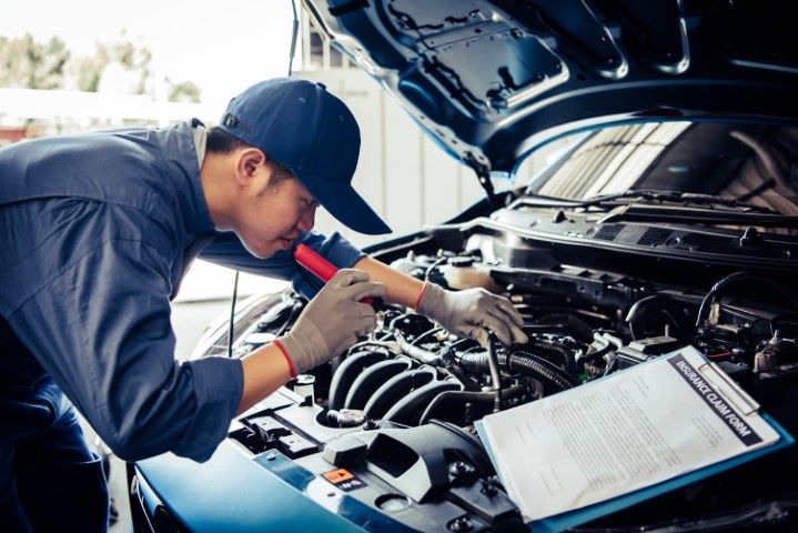 Mechanic in blue uniform inspects engine with flashlight, clipboard nearby, in a garage.