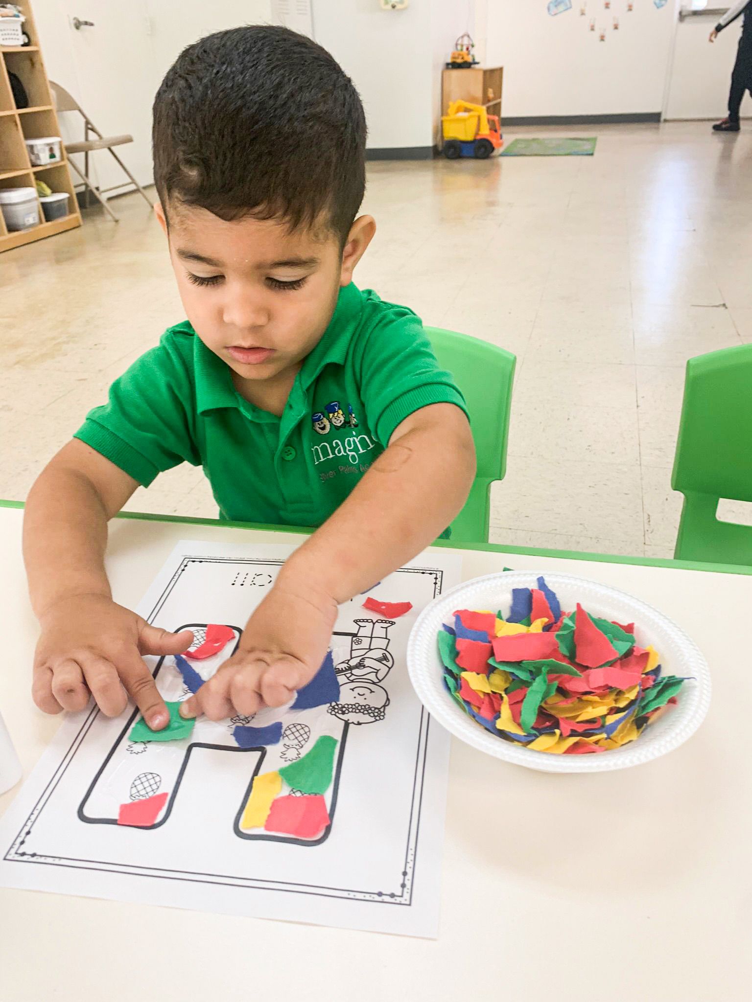 A young boy in a green shirt is sitting at a table playing with a bowl of stickers.