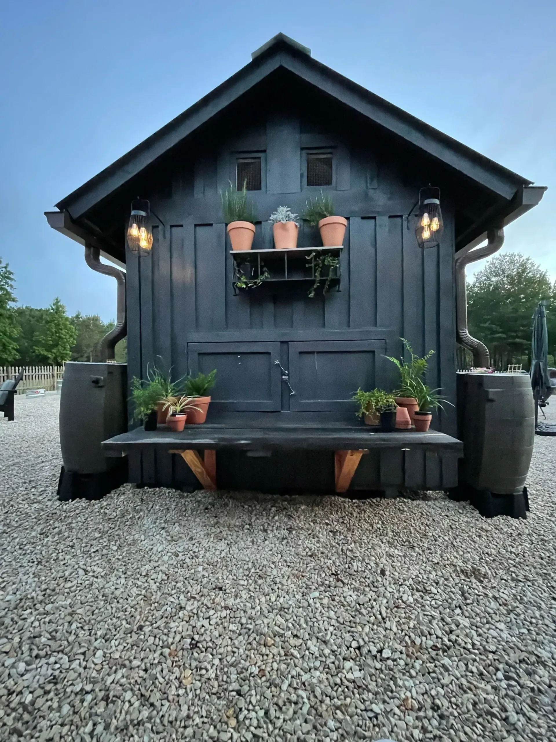 Black shed with bench and potted plants. Gravel ground, two rain barrels, and two sconces.