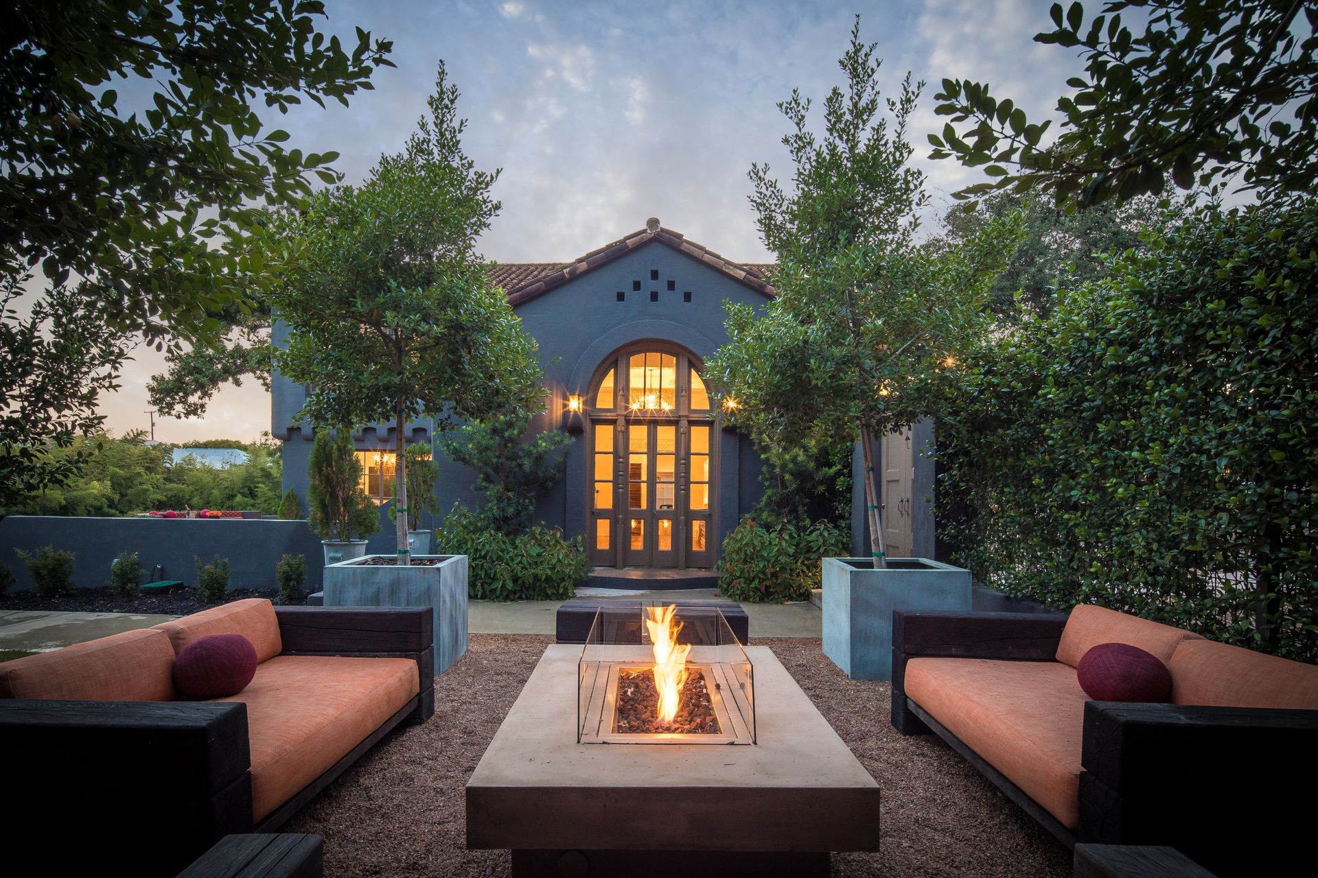 Outdoor patio with fire pit, orange sofas, and a building with arched doorway at dusk.